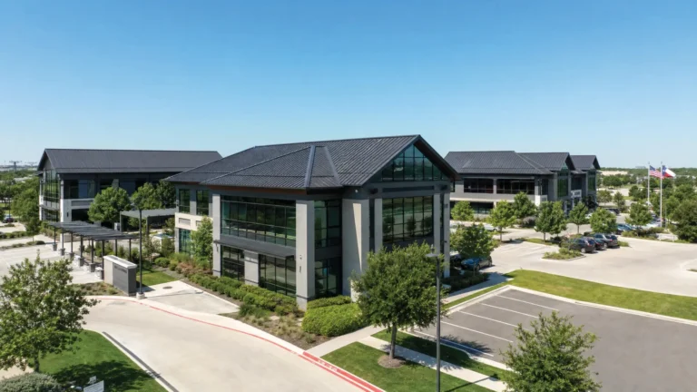 Aerial view of modern office buildings with black roofs, large glass windows, and surrounding trees. An empty parking lot and driveways are visible under a clear blue sky. Three American flags are in the background.